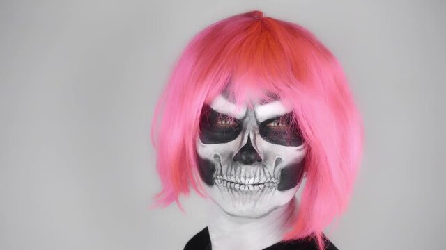 Cheerful funny man in halloween skeleton makeup in a pink wig turns and flaunts in front of the camera. Shooting in the studio. Gray background