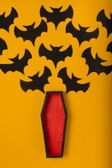 an open coffin with red padding from which black handmade paper bats fly out. halloween concept on yellow background
