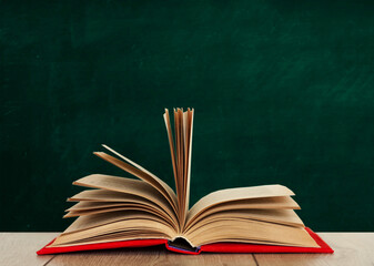 Open book textbook in a red cover on a wooden desk on the background of a green school chalkboard. Back to school distance home education.Quarantine concept of stay home.