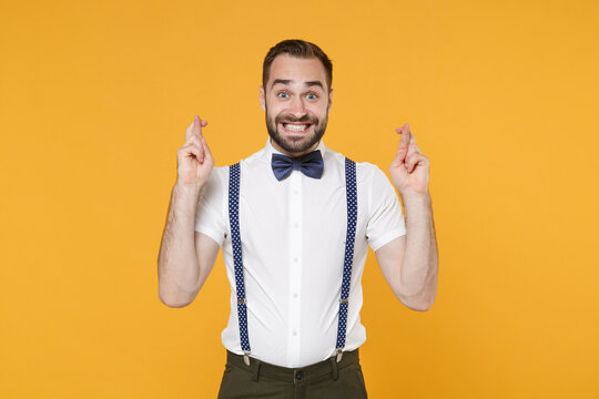 Excited young bearded man 20s wearing white shirt bow-tie suspender posing waiting for special moment keeping fingers crossed making wish isolated on yellow color wall background, studio portrait.