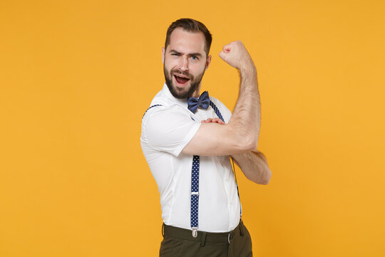 Amazed cheerful strong young bearded man 20s wearing white shirt bow-tie suspender posing showing biceps muscles keeping mouth open isolated on bright yellow color wall background, studio portrait.