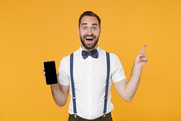 Excited young bearded man 20s in white shirt bow-tie suspender pointing index finger up hold mobile cell phone with blank empty screen mock up copy space isolated on yellow background studio portrait.