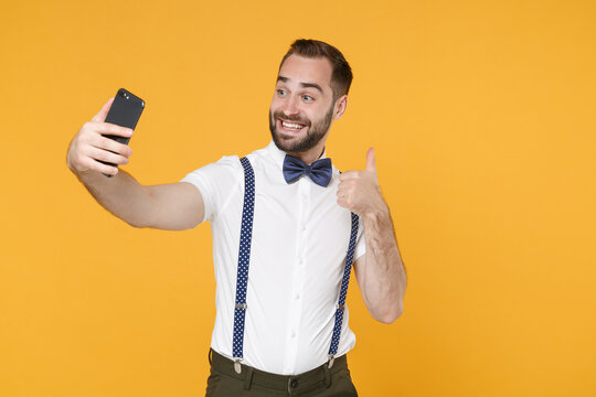 Smiling funny young bearded man 20s in white shirt bow-tie suspender posing doing selfie shot on mobile phone showing thumb up isolated on bright yellow color wall background studio portrait.