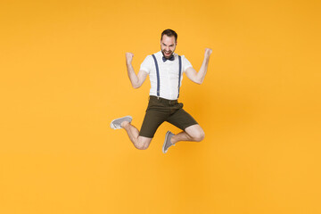 Fototapeta na wymiar Full length portrait of happy young bearded man 20s wearing white shirt suspender shorts posing jumping doing winner gesture looking camera isolated on bright yellow color wall background studio.