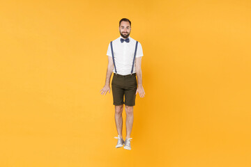 Fototapeta na wymiar Full length portrait of smiling funny young bearded man 20s wearing white shirt suspender shorts posing jumping having fun looking camera isolated on bright yellow color wall background studio.
