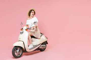 Fototapeta na wymiar Full length portrait of shocked young brunette woman wearing white summer clothes hat glasses keeping mouth open looking aside sitting driving moped isolated on pastel pink colour background studio.