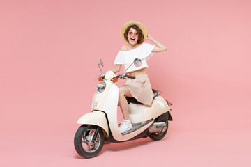 Fototapeta na wymiar Full length portrait of surprised cheerful young brunette woman 20s wearing white summer clothes hat glasses keeping mouth open sitting driving moped isolated on pastel pink colour background studio.