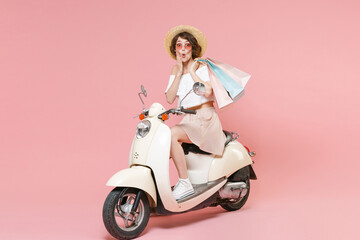 Fototapeta na wymiar Full length portrait of amazed young woman in white clothes hat glasses hold package bag with purchases after shopping put hand on cheek driving moped isolated on pastel pink colour background studio.