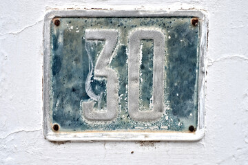 Number 30, thirty, a weathered blue plate on a white wall background.
