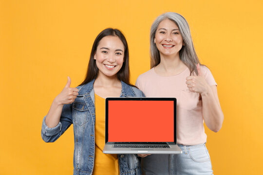 Smiling family asian women girls mother daughter in casual clothes hold laptop pc computer with blank empty screen mock up copy space showing thumbs up isolated on yellow background studio portrait.