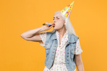 Fototapeta na wymiar Funny elderly gray-haired female woman 60s 70s wearing casual dress denim waistcoat birthday hat celebrating blowing in pipe looking aside isolated on yellow color wall background studio portrait.