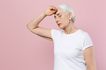 Exhausted tired elderly gray-haired female woman 60s 70s wearing white blank design casual t-shirt posing put hand on head keeping eyes closed isolated on pastel pink color background studio portrait.