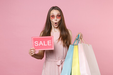 Fototapeta premium Shocked young brunette woman in pink summer dotted dress glasses hold package bag with purchases after shopping sign with SALE title looking camera isolated on pastel pink background studio portrait.