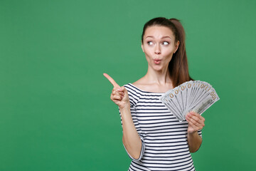 Shocked young brunette woman 20s in striped casual clothes hold fan of cash money in dollar banknotes pointing index finger aside on mock up copy space isolated on green background studio portrait.