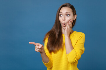 Shocked young brunette woman 20s wearing yellow casual clothes posing standing pointing index finger aside on mock up copy space put hand on cheek isolated on blue color background studio portrait.