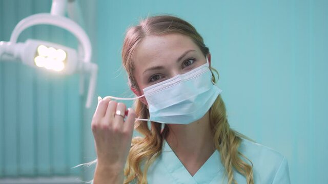 Young pretty woman dentist takes off her protective face mask and looks into the camera smiling. Close-up.