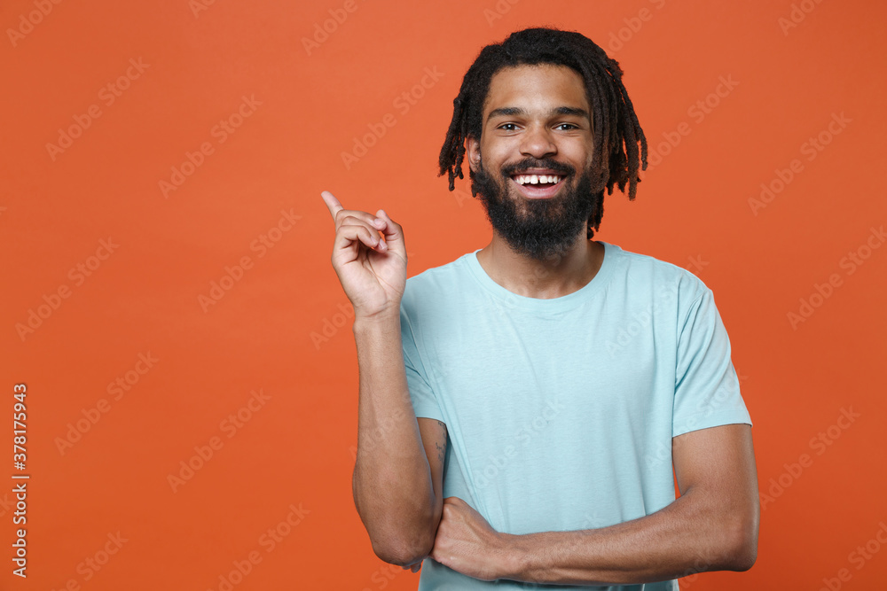 Wall mural Funny young african american man guy wearing blue casual t-shirt posing isolated on orange background studio portrait. People emotions lifestyle concept. Mock up copy space. Pointing index finger up.