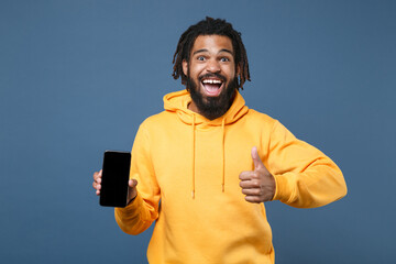Fototapeta premium Excited young african american man guy in yellow hoodie isolated on blue background studio. People lifestyle concept. Mock up copy space. Hold mobile phone with blank empty screen showing thumb up.