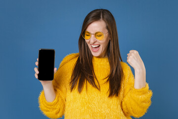 Happy young brunette woman 20s in yellow fur sweater eyeglasses hold mobile phone with blank empty screen mock up copy space doing winner gesture isolated on blue color background studio portrait.