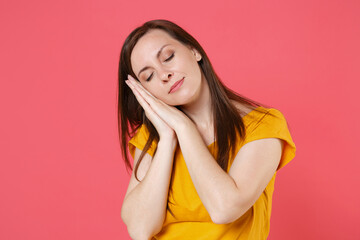 Relaxed beautiful attractive young brunette woman 20s wearing yellow casual t-shirt posing sleeping with folded hands under cheek keeping eyes closed isolated on pink color background studio portrait.