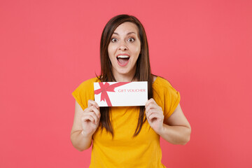 Excited surprised young brunette woman 20s in yellow casual t-shirt posing holding in hands gift certificate keeping mouth open looking camera isolated on pink color wall background studio portrait.