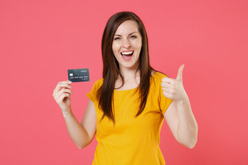 Cheerful young brunette woman 20s in yellow casual t-shirt posing standing holding in hands credit bank card showing thumb up looking camera isolated on pink color wall background studio portrait.