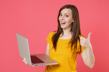 Funny cheerful young brunette woman 20s wearing yellow casual t-shirt posing and working on laptop pc computer showing thumb up looking camera isolated on pink color wall background studio portrait.