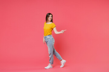 Fototapeta na wymiar Full length portrait side view of smiling beautiful young brunette woman 20s wearing yellow casual t-shirt posing standing pointing index finger aside isolated on pink color wall background studio.