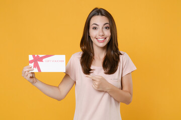 Fototapeta na wymiar Smiling pretty young brunette woman 20s wearing pastel pink casual t-shirt posing pointing index finger on gift certificate looking camera isolated on bright yellow color background studio portrait.