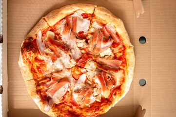 Delicious pizza with cheese and prosciutto in a cardboard box. Pizza delivered to the customer. Close-up of pizza cut into pieces