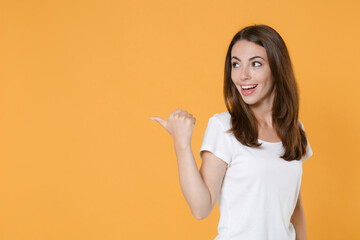 Side view of cheerful smiling funny young brunette woman 20s in white blank empty design casual t-shirt pointing thumb aside on mock up copy space isolated on yellow color background studio portrait.