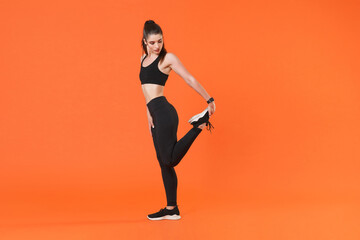 Fototapeta na wymiar Full length side view portrait young fitness sporty woman in black sportswear posing training working out doing stretching exercising for legs looking down isolated on orange color background studio.
