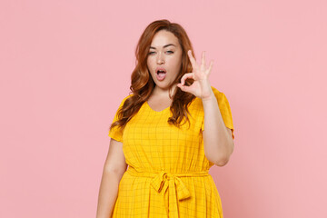 Stunning attractive shocked young redhead plus size body positive female woman girl in yellow dress posing showing OK gesture looking camera isolated on pastel pink color background studio portrait.