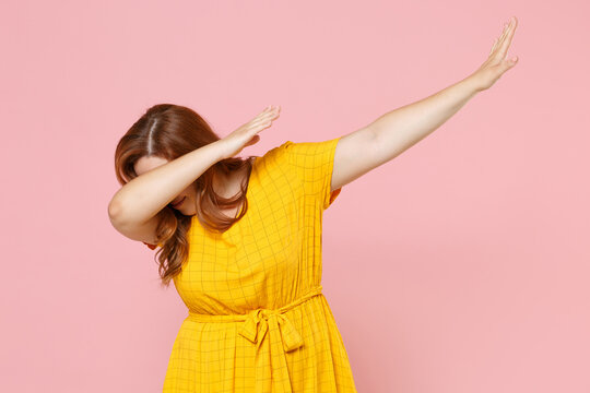 Young redhead plus size body positive female woman in yellow dress doing dab dance gesture hip hop hands youth sign cover hiding covering face isolated on pastel pink color background studio portrait.