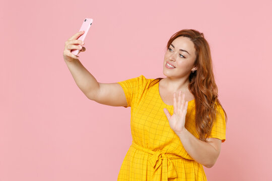 Smiling young redhead plus size body positive female woman girl in yellow dress posing doing selfie shot on mobile phone waving greeting with hand isolated on pink color background studio portrait.