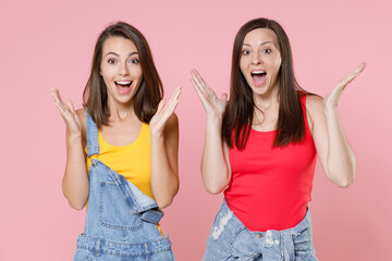 Two excited surprised beautiful attractive young women friends 20s in casual denim clothes spreading hands keeping mouth open looking camera isolated on pastel pink colour background, studio portrait.