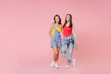 Full length portrait of two smiling cheerful attractive young brunette women friends 20s wearing casual denim clothes posing hugging looking camera isolated on pastel pink colour background in studio.