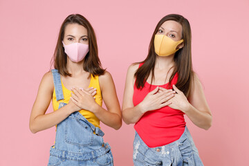 Two young women friends 20s in casual denim clothes sterile face mask to safe from coronavirus virus covid-19 holding hands folded on hearts isolated on pastel pink colour background studio portrait.