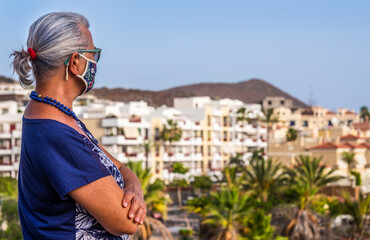Fototapeta na wymiar Portrait of senior woman with flower mask because of coronavirus walking outdoors at sunset, looking at landscape - concept of active elderly during retirement