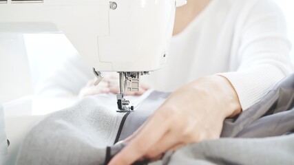 Female hands sew on a white sewing machine close-up. Concept of sewing in modern bright studio, woman in white sweater sewing grey cloth in process, selective focus