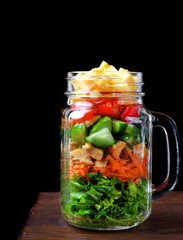 Layered salad with cheese, kale, carrot, chicken, cucumbers and cherry tomatoes in a glass mason jar against the black background