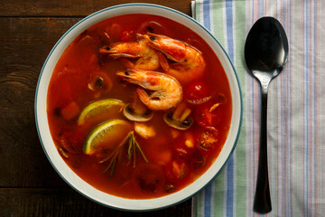 Tom yam soup with shrimp and lime in a plate next to a spoon on a napkin.