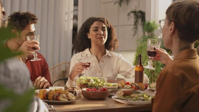 Back view of mixed-race curly-haired young woman sitting at dining table with friends, holding wine glass in her hand, making toast and then clinking glasses with the othersBack view of mixed-race cur