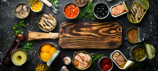 Canned vegetables, beans, fish and fruits in tin cans on black stone background. Food stocks.