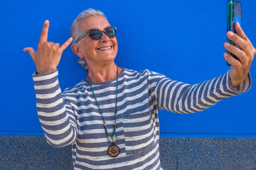 Naklejka premium Cheerful attractive senior woman with white hair smiling happy making a selfie using smart phone standing - blue background - tech and social elderly people