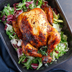 Top view of whole roasted chicken with fresh salad in black dish.