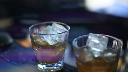 Two glasses with whiskey and ice on bar counter. Close up shot of alcohol in bar, blurred background. Two cocktails with ice ready to be served