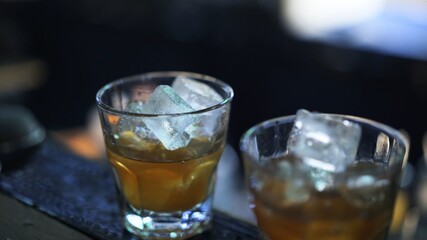 Two glasses with whiskey and ice on bar counter. Close up shot of alcohol in bar, blurred background. Two cocktails with ice ready to be served