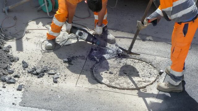 Two workers in protective orange uniform using heavy drilling electric hammer for breaking concrete on the road, hard working on construction site for opening clogged sewerage on the street