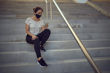 Girl with black mask watching mobile phone sitting on some stairs.